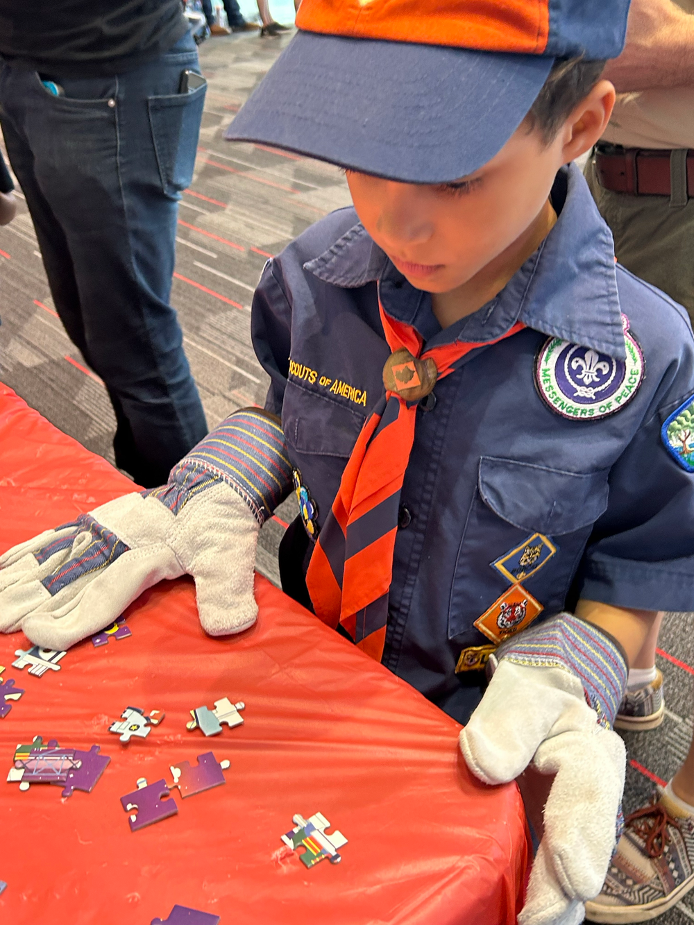 A young boy wearing garden gloves, a blue polo with an orange tie, and a blue and orange hat looks down at a table containing puzzle pieces.